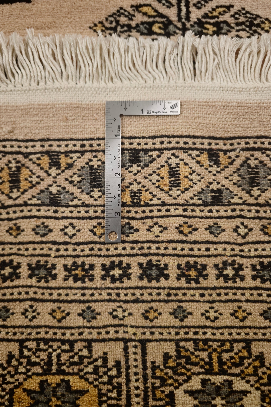 Close-up of a patterned rug with a metal L-square ruler placed vertically to show the scale of the woven design and fringe.